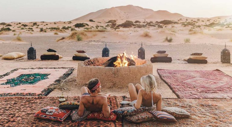 Two influencers looking the Sahara desert of Merzouga in Morocco around the firepit in a camp.